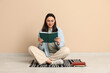© Pixel-Shot - Young woman sitting on floor and reading book near beige wall