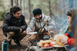 © qunica.com - Two friends cook and smile around a small campfire in a wooded campsite. A group enjoys outdoor cooking and conversation during a chilly camping trip.