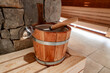 © Aerial Film Studio - A wooden sauna bucket with metal bands and a ladle rests on a timber bench by a stacked stone wall. Warm amber light and shallow depth of field suggest a luxury spa setting.