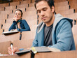 © Connect Images - Two students in a lecture hall with notebooks, sitting among rows of wooden seats. Germany