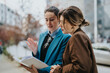 © qunica.com - Two women in business attire review a notebook on a city street, engaged in collaboration and discussion.