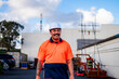 © NZstockphoto - A smiling construction worker in a bright orange uniform walks through an outdoor job site