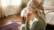 © 24DIY - A person ties a floral headscarf revealing a bald head while sitting on the floor near burning incense and a mat