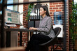 © DC Studio - Asian businesswoman examines financial data on her desktop monitor in brick wall workspace. Female entrepreneur analyzes business performance reports on computer, near an office window.