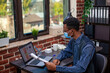 © DC Studio - Startup owner with eyeglasses carefully reviewing sales data on laptop, holding clipboard filled with marketing charts in office. Manager wearing earbuds and face mask, working on business project.