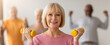 © Prostock-studio - A group of elderly individuals participates in a fitness class. A woman in front lifts yellow weights and smiles while the others are exercising in the background. The setting is a well-lit room.