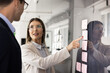 © fizkes - Young businesswoman engaged in discussion with male colleague while pointing at glass wall covered in colorful sticky notes, explaining strategy or insights, mapping out tasks. Brainstorming, planning