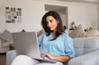 © fizkes - Focused young woman reviewing products or making purchases online using laptop seated on cozy sofa in living room, checking bank accounts, managing freelance assignments, scrolling social media news