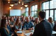 © miss irine - People sit at tables in a modern room, listening to a speaker. Brick walls and large windows create an industrial loft atmosphere. Attendees focus intently on the presentation happening before them.
