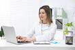 © New Africa - Smiling doctor in scrubs working with laptop at white table in clinic