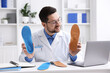 © New Africa - Smiling orthopedist in glasses and medical coat with foot insoles at white desk in clinic
