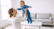 © NeuroCake - Happy mother playing with her laughing baby boy in the living room. Mom lifting her infant son in the air at home. Joyful family and motherhood concept