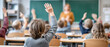 © Kowit - Children in a classroom raise their hands to participate while a teacher stands at the front near a chalkboard.