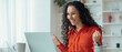 © Prostock-studio - A woman with curly hair expresses joy while sitting at a desk. She is looking at her laptop and has her hands raised in celebration. The bright room has plants and organized items.