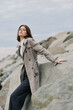 © SHOTPRIME STUDIO - Young woman in long coat poses on rocky shore under cloudy sky. Fashion model leans on stone with confident look. Concept for autumn style and natural landscape.