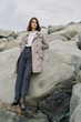 © SHOTPRIME STUDIO - Young woman in stylish coat and dark trousers stands confidently on large rocks outdoors. Overcast sky and natural environment create a calm, contemplative mood in this fashion portrait.