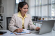 © apichat - Asian businesswoman working on laptop, taking notes at office