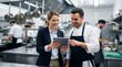 © NORN - Culinary Collaboration: A chef and a manager engage in a discussion, sharing ideas and expertise in a professional kitchen setting, using a tablet to streamline the process.