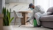 © standret - Male pest control technician in protective suit sprays insecticide on floor in modern living room with dining area and green plant in the foreground