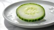 © SERHII - Cucumber slice drop single round fresh vegetable white porcelain plate dramatic side lighting creating strong shadows water droplets edges minimalist kitchen clean surfaces motion