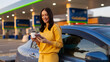 © Home-stock - Roadside digital break. Happy woman drinking coffee and looking at cellphone, standing by her car at gas station, panorama shot