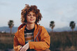 © SHOTPRIME STUDIO - Smiling woman with curly hair wearing a rainbow sweater and orange jacket sitting outdoors near palm trees. The woman holds a smartphone, representing lifestyle and casual fashion.