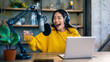 © Iryna - Young asian female adult podcaster smiling with headphones microphone laptop in cozy home studio wearing yellow sweater holding drink while recording