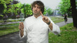 © Krakenimages.com - Man chef wearing white coat holding whisk in left hand points finger with bare hand toward viewer on street by tram tracks; confidence humor.