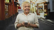 © Krakenimages.com - Senior woman holding smartphone and tapping screen with hands visible, standing on a narrow street lined with shops and awnings, looking upward; curiosity.