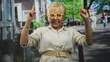 © Krakenimages.com - Senior hispanic woman with yellow glasses raises both arms and points fingers upward on a sunny street with buildings and trees; joy celebration empowerment.