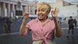 © Krakenimages.com - Senior woman in striped shirt pointing index finger and making ok sign with hand while smiling on a busy street near large columns; joy travel.