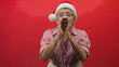 © Krakenimages.com - Woman in santa hat and glasses cupping hands around mouth while calling out in a studio with vivid red backdrop and striped shirt; holiday joy.