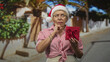 © Krakenimages.com - Senior woman wearing a santa hat and sweater, smiling while holding a wrapped gift and greeting in a studio; holiday joy warmth.