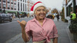 © Krakenimages.com - Senior woman wearing a santa hat and striped tied shirt points thumb over shoulder and smiles on a busy shopping street; holiday joy.