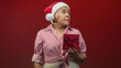 © Krakenimages.com - Woman wearing santa hat holding red gift box with hand on abdomen, looking up in studio against red backdrop; holiday nostalgia.