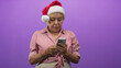 © Krakenimages.com - Woman in santa hat taps smartphone with hands against purple studio backdrop wearing striped shirt and bracelets; quiet contemplation.