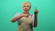 © Krakenimages.com - Woman raising middle finger and pointing at it in a green studio, senior hispanic wearing a beige apron with rings and bracelets, smiling broadly; defiance.