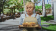 © Krakenimages.com - Senior woman in apron holding a wooden tray of muffins, hands forward presenting baked goods with neutral gaze amid trees in a forest; determination.