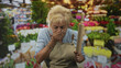 © Krakenimages.com - Senior hispanic woman cook wearing beige apron, holding a rolling pin and covering her mouth with hand, eyes closed, standing at a flower market stall; disgust concern.