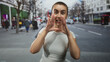 © Krakenimages.com - Woman with shaved hair cups hands around mouth shouting on busy urban street by red bus and pedestrians; urgent alert.