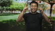 © Krakenimages.com - Young chinese man in black shirt points index finger upward in sunlit park with green trees; optimism aspiration.