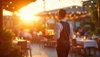 © miss irine - Waiter brings beer to outdoor restaurant tables at sunset. People dine on patio with string lights ambiance. Evening meal service with golden hour lighting.