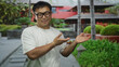 © Krakenimages.com - Young chinese man wearing glasses and a white t shirt showing open palms in front of a building facade; warm welcome.