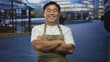 © Krakenimages.com - Young chinese man wearing apron crossing arms on street in city center while smiling broadly; confidence.