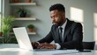 © Viktor - A Black man in a suit types on a laptop in a bright office with plants. He looks focused on his work, sitting at a desk, with sunlight streaming in.