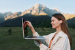 © SHOTPRIME STUDIO - A cheerful woman works outdoors with a laptop in a scenic field beneath mountain peaks. She smiles while typing, conveying focus, productivity, and a refreshing outdoor work vibe.