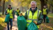 © miss irine - Man with gray hair smiles wearing yellow vest holding green bags. People clean park in autumn. Volunteers work together, collecting rubbish for eco initiative. Team supports environment, community.