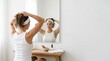 © lilPENtacle - Woman Looking in Mirror Fixing Hair with Headband at Minimalist Vanity. Morning Skincare Routine, Clean Beauty Prep, and Self Care with White Copy Space.