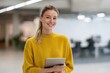 © Andrii Zastrozhnov - Young caucasian female smiling in office holding tablet wearing yellow sweater confident professional environment coworker blurred background