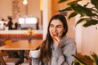 © zvkate - Happy young woman in casual grey hoodie holding white cup and eating delicious cookie or biscuit while looking away with cheerful smile sitting in cozy cafe interior during morning break time